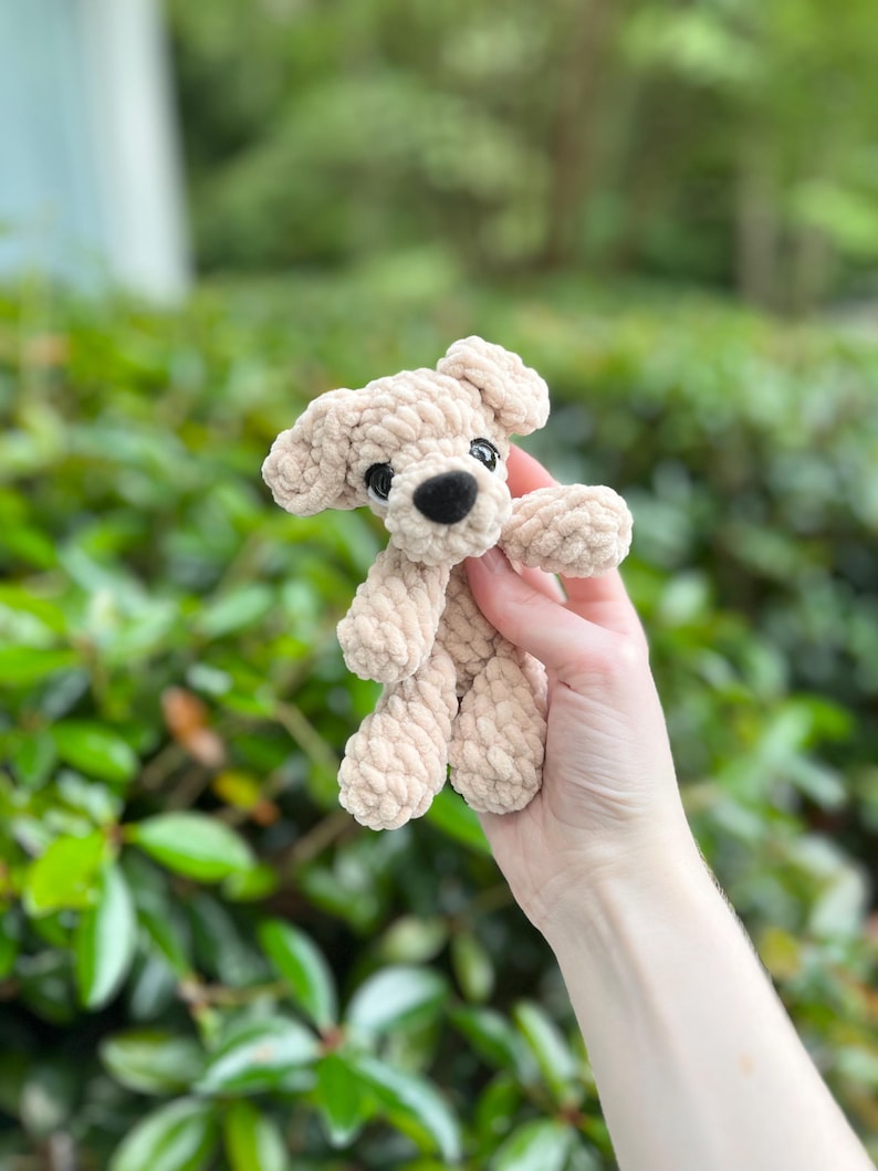 以下が含まれることがあります： 厚みのある質感の糸で編まれた、ベージュ色の小さなかぎ針編みの犬のぬいぐるみ。このぬいぐるみには、垂れ下がった耳が2つ、黒い鼻が付いた丸みを帯びた鼻先、そして2つの小さくて暗い丸い目があります。
