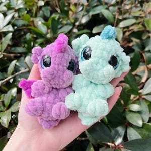 May include: Two handmade crochet plush toys, one lavender and one mint green, are held in a hand. The lavender toy has a pink horn and wings, while the mint green toy has a blue tuft. Both have large, shiny black eyes.