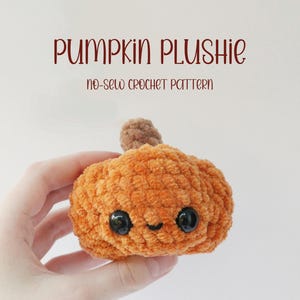 Peut inclure: Un ourson en crochet en forme de citrouille avec une tige brune et des yeux noirs. L'ourson est tenu dans une main et a un visage souriant. Le texte "Pumpkin Plushie no-sew crochet pattern" est au-dessus de l'ourson.