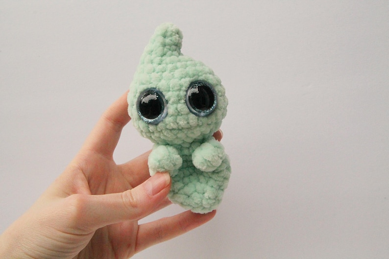 Puede incluir: Un juguete de peluche fantasma verde tejido a crochet con grandes ojos brillantes.