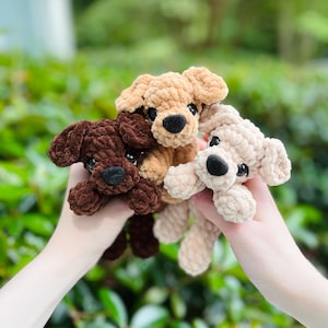 以下が含まれることがあります： ダークブラウン、ミディアムタン、ライトクリームの色合いの柔らかく質感のある糸で作られた3匹の小さな犬のぬいぐるみ。それぞれのぬいぐるみには、垂れ下がった耳、丸い黒い鼻、暗くて光沢のある目があり、太いかぎ針編みのステッチで仕上げられています。