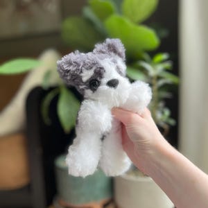 May include: A small, plush toy dog with a black nose and eyes, and gray and white fur. The toy dog is held up in front of a blurred background of plants and pots.