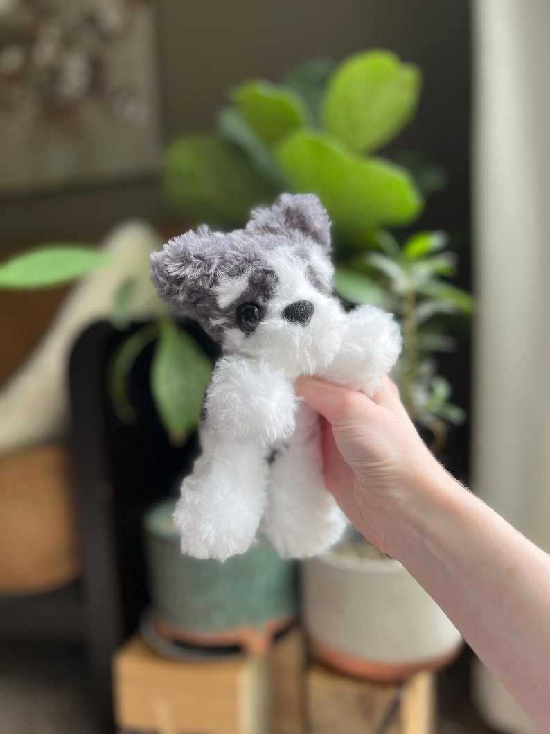 May include: A small, plush toy dog with a black nose and eyes, and gray and white fur. The toy dog is held up in front of a blurred background of plants and pots.
