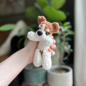 May include: A handmade crochet dog toy in white and brown. The dog has a white body, brown ears, and a brown spot around one eye. The toy is held in a person's hand.