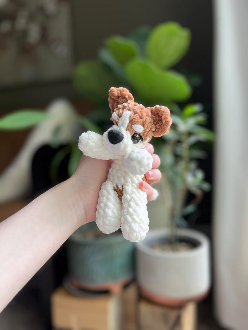 May include: A handmade crochet dog toy in white and brown. The dog has a white body, brown ears, and a brown spot around one eye. The toy is held in a person's hand.