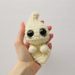 Puede incluir: Un juguete de peluche de fantasma tejido a crochet con grandes ojos saltones. El fantasma es blanco y tiene una forma redondeada.
