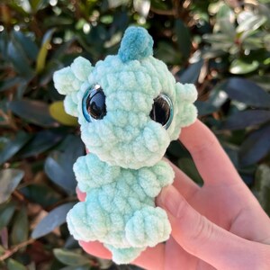 May include: A light teal crocheted creature with large, reflective black eyes and a small, teal fin on its head. The plush toy has a textured, bumpy surface and small arms and legs. The background is blurred green foliage.