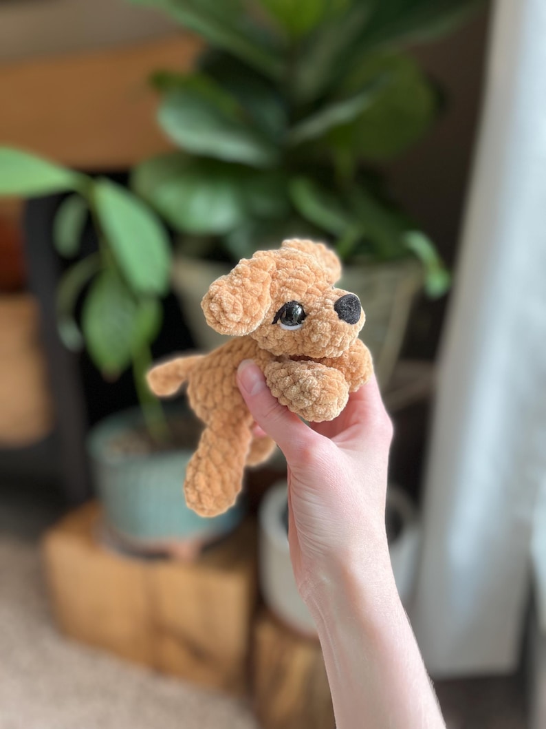 以下が含まれることがあります： 柔らかい質感の糸で作られた、小さなライトブラウンの犬のぬいぐるみ。このぬいぐるみは、垂れ耳、小さな黒い鼻、暗くて光沢のあるプラスチック製の目が特徴です。シンプルな丸みを帯びた体型と短い手足でデザインされています。