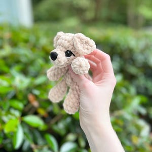 以下が含まれることがあります： 柔らかく太い糸で編まれた、ベージュ色の小さなかぎ針編みの犬のぬいぐるみ。垂れ耳、丸い黒い鼻、暗く光沢のあるプラスチック製の目が特徴です。糸の質感がフィギュア全体にはっきりと見て取れます。