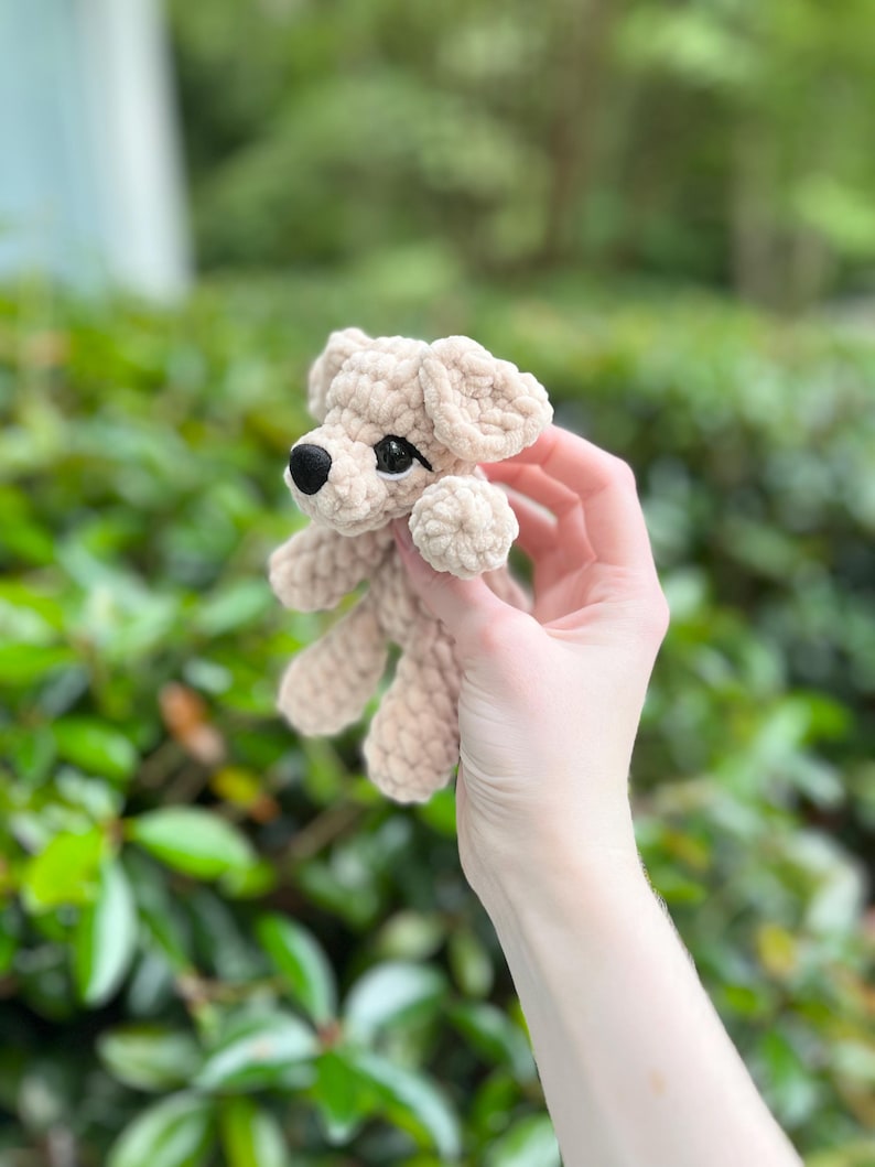 以下が含まれることがあります： 柔らかく太い糸で編まれた、ベージュ色の小さなかぎ針編みの犬のぬいぐるみ。垂れ耳、丸い黒い鼻、暗く光沢のあるプラスチック製の目が特徴です。糸の質感がフィギュア全体にはっきりと見て取れます。