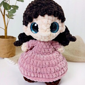 May include: A handmade crochet doll with a light pink dress and dark brown hair. The doll has large, sparkly blue eyes and brown shoes. The doll is made of soft, textured yarn and is a charming, whimsical creation.