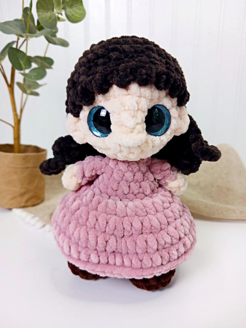 May include: A handmade crochet doll with a light pink dress and dark brown hair. The doll has large, sparkly blue eyes and brown shoes. The doll is made of soft, textured yarn and is a charming, whimsical creation.