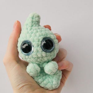 Puede incluir: Un peque&ntilde;o juguete de peluche tejido a crochet de color verde claro con grandes ojos azules brillantes. El juguete tiene un cuerpo redondeado y una cabeza puntiaguda.