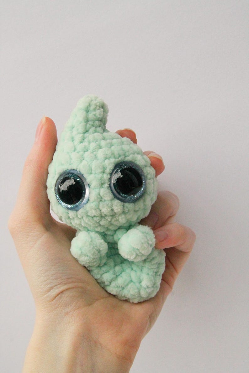Puede incluir: Un peque&ntilde;o juguete de peluche tejido a crochet de color verde claro con grandes ojos azules brillantes. El juguete tiene un cuerpo redondeado y una cabeza puntiaguda.