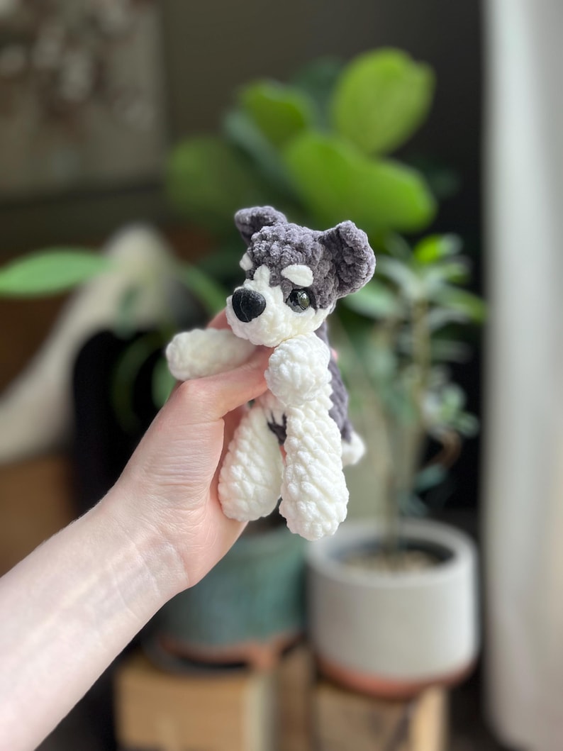 May include: A handmade crochet dog toy in gray and white. The dog has a black nose and eyes, and white paws. The toy is held in a person's hand, with a blurred background of plants and a pot.