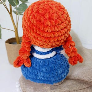 May include: A handmade plush doll with bright orange hair styled in braids. The doll wears a blue dress with white accents. The doll is made of a soft, textured material, and is a charming handmade item.