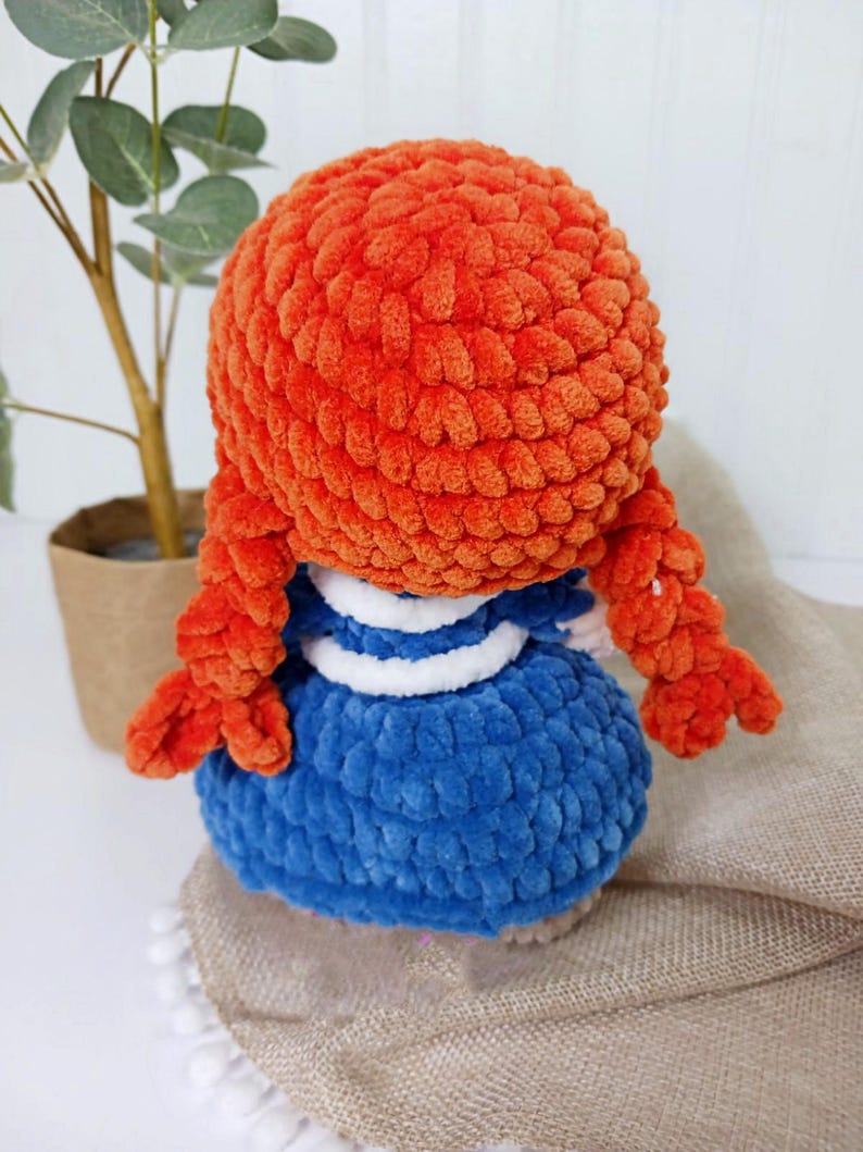 May include: A handmade plush doll with bright orange hair styled in braids. The doll wears a blue dress with white accents. The doll is made of a soft, textured material, and is a charming handmade item.