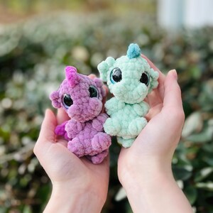 May include: Two small, handmade crochet dragon toys. One is purple with a pink crest, the other is mint green with a blue crest. Both have large, black-pupiled eyes and are held in open hands.