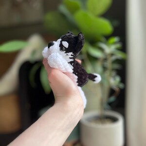 May include: A handmade crochet dog in black and white. The small stuffed animal has a black head and back with white accents on the face, chest, and paws. The dog is lying down, with its paws tucked in. The background is blurred, with green plants visible.