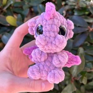 May include: A handmade, lavender-colored crochet dragon with large, reflective black eyes. The dragon has small, magenta wings and a matching crest. The plush toy is soft and textured, perfect for a child's room or as a unique gift.