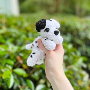 以下が含まれることがあります： ダルメシアンの子犬の形をした、手編みの小さなかぎ針編みのぬいぐるみ。本体は白を基調とし、黒い斑点、黒い耳、黒い鼻が特徴です。暗く光沢のあるプラスチック製の目と、柔らかい質感の毛糸で作られた構造をしています。