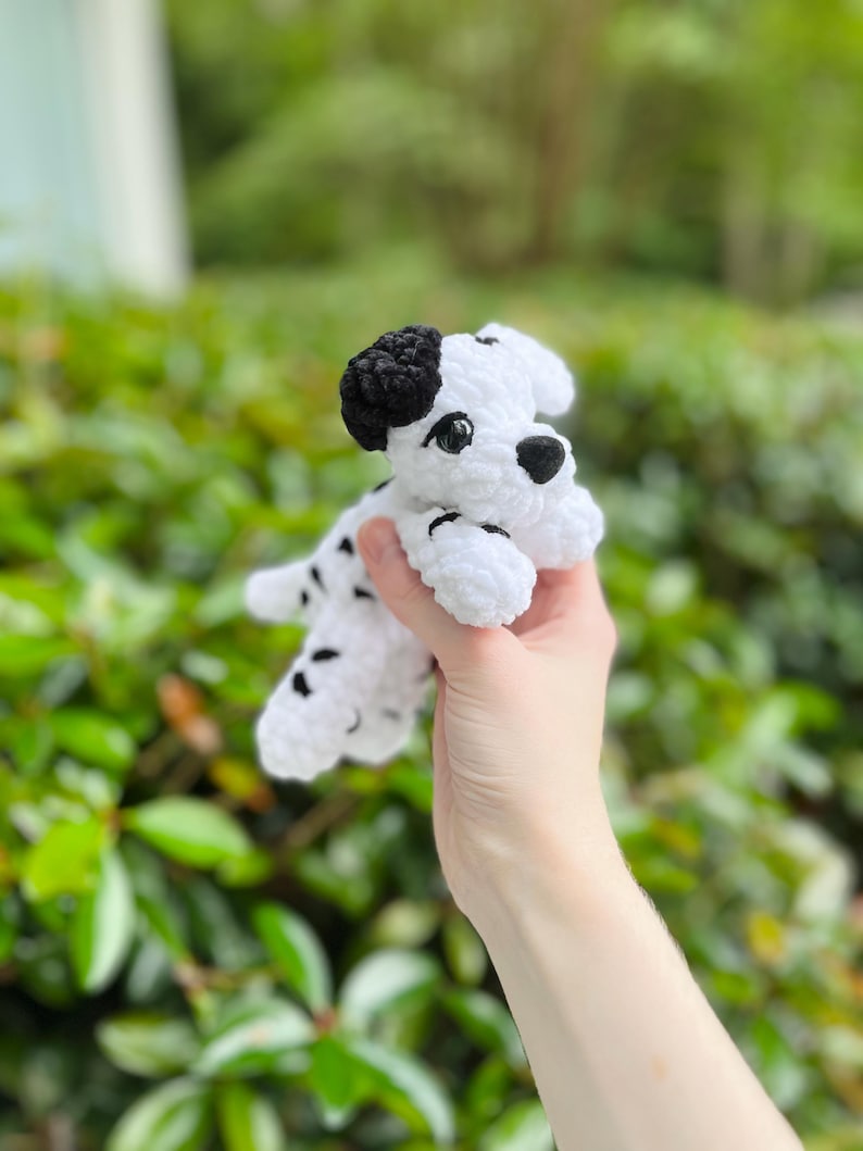 以下が含まれることがあります： ダルメシアンの子犬の形をした、手編みの小さなかぎ針編みのぬいぐるみ。本体は白を基調とし、黒い斑点、黒い耳、黒い鼻が特徴です。暗く光沢のあるプラスチック製の目と、柔らかい質感の毛糸で作られた構造をしています。