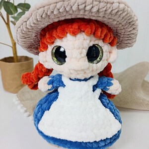 May include: A handmade crochet doll with red hair in pigtails, wearing a blue dress with a white apron and a beige hat. The doll has large, glittery eyes and is made of soft, plush yarn.