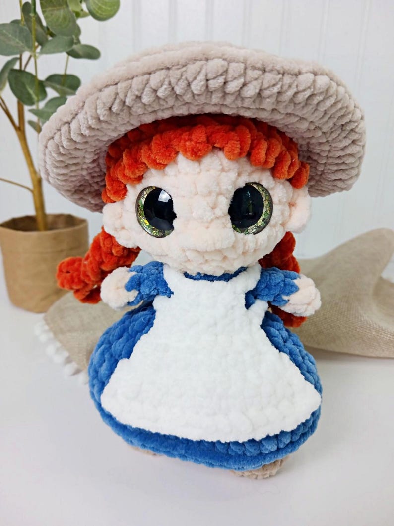 May include: A handmade crochet doll with red hair in pigtails, wearing a blue dress with a white apron and a beige hat. The doll has large, glittery eyes and is made of soft, plush yarn.