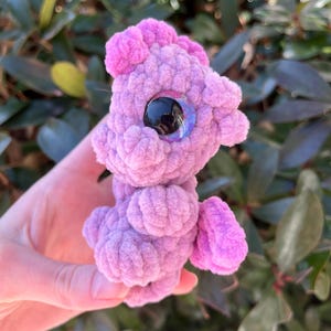 May include: A handmade, lavender-colored crochet creature with a large, iridescent eye. The plush toy has a textured, fuzzy appearance and features small, rounded appendages. The background is blurred, with green foliage.