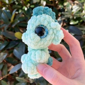 May include: A handmade, light teal crochet seahorse with a textured, plush appearance. The seahorse has a darker teal mane and tail, and large, expressive blue and black eyes. The plush toy is held against a backdrop of green foliage.
