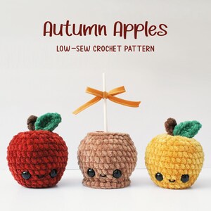Pode incluir: Três maçãs de crochê, uma vermelha, uma marrom e uma amarela, com folhas verdes e rostos sorridentes. A maçã marrom está em um palito com uma fita amarela.