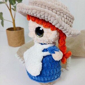 May include: A handmade plush doll with a light brown hat, red hair, and a blue dress. The doll has large, glittery eyes and a white apron. The doll is approximately 6 inches tall and is a charming, handcrafted item.