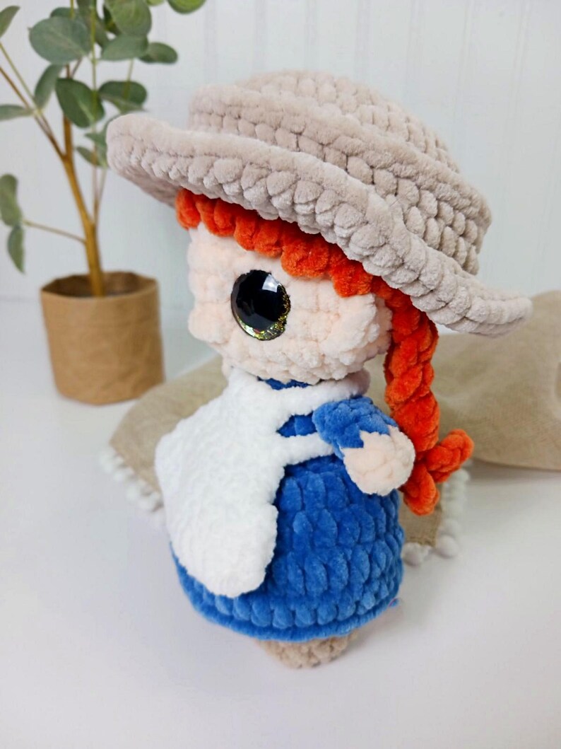 May include: A handmade plush doll with a light brown hat, red hair, and a blue dress. The doll has large, glittery eyes and a white apron. The doll is approximately 6 inches tall and is a charming, handcrafted item.