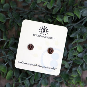 May include: A pair of wooden stud earrings with a transgender symbol engraved on each. The earrings are displayed on a white card with the text "Beyond Our Story; Don't ever be afraid to show your true colors!"