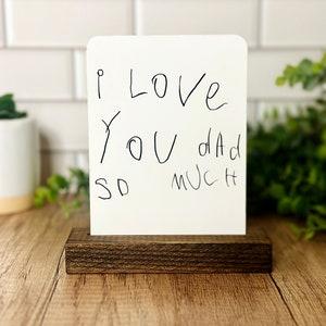 May include: A white card with the handwritten message "I LOVE YOU Dad SO MUCH" is displayed in a dark brown wooden block. The card is centered and the wooden block is rectangular. The background is a light-colored wood surface.