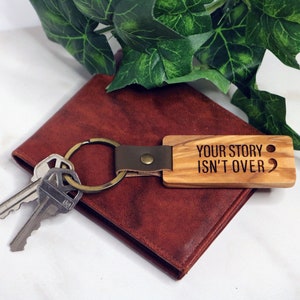 May include: Wooden keychain with a brown leather strap and a metal ring. The keychain has the text "YOUR STORY ISN'T OVER" engraved on it, along with a semicolon.
