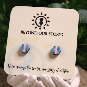 May include: A pair of wooden earrings with a hexagonal shape. The earrings are painted with blue, white, and pink stripes. The earrings are displayed on a white card with the text "BEYOND OUR STORY; Help change the world, one story at a time."