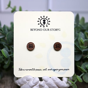 Op de afbeelding: Een paar houten oorbellen met een eenvoudig ontwerp. De oorbellen zijn bruin en hebben een zwart puntkomma-symbool erop. De oorbellen worden getoond op een witte kaart met de tekst "Beyond Our Story;" en "Take a moment to pause, rest, and regain your peace."