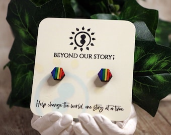 Puede incluir: Pendientes de botón hexagonales con diseño de arcoíris. Se muestran en una tarjeta con el texto "BEYOND OUR STORY;" y "Help change the world, one story at a time."