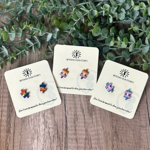 May include: Three pairs of small wooden puzzle piece earrings, each pair featuring a different pride flag design. The earrings are displayed on white cards with the text "Beyond Our Story: Don't ever be afraid to show your true colors!"
