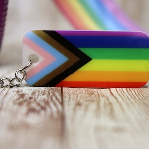 May include: A rainbow pride flag keychain with a black and brown chevron on a white background. The keychain is made of plastic and has a silver metal chain.