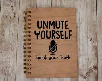 May include: A wooden spiral bound notebook with the text "Unmute Yourself" and a black and white microphone icon with a line through it. The text "Speak your truth" is below the icon.