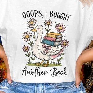 Puede incluir: Camiseta blanca con un diseño caprichoso de un pato llevando libros, rodeado de flores. El texto dice "OOOPS, I BOUGHT Another Book". El diseño utiliza colores pastel suaves, creando una estética encantadora y juguetona.