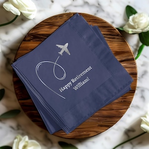 May include: Navy blue napkins with a silver airplane design and the text "Happy Retirement William!". The napkins are stacked on a wooden plate, surrounded by white roses.