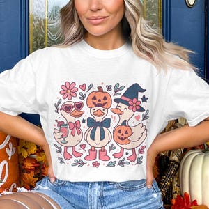 Op de afbeelding: Wit T-shirt met een Halloween-ontwerp. De afbeelding toont drie cartoon eenden met pompoenhoofden, heksenhoeden en zonnebrillen. De eenden zijn omringd door bloemen en harten, wat een feestelijke en speelse uitstraling creëert.