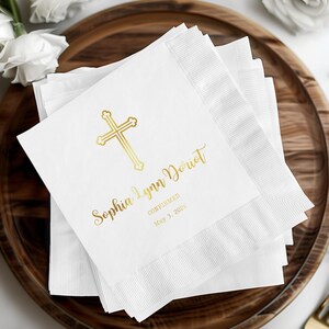 May include: White paper napkins with a gold cross design and custom text. The napkins are stacked on a wooden tray. The text reads "Sophia Lynn Doriot Confirmed May 3, 2026".