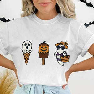 Puede incluir: Camiseta blanca con tres diseños de helados de Halloween. Los diseños incluyen un fantasma, un helado con forma de calabaza y un fantasma con gafas de sol y traje de baño sosteniendo una cesta. La camiseta es informal.