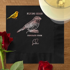 May include: A black cocktail napkin with a bird illustration and the text "FLYING HIGH!" and "CONGRATS FROM Sadie". A yellow bird is on the left. A glass of wine and red roses are in the background.