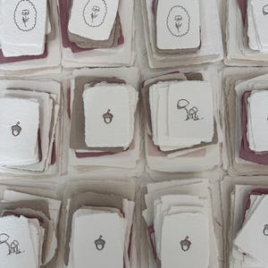 May include: Multiple stacks of rectangular handmade paper with deckled edges in shades of white, cream, mauve, and taupe. Each stack features a top sheet printed with black ink illustrations of flowers, acorns, or mushrooms within simple borders.