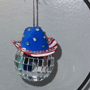 May include: A silver disco ball bauble with a blue and red cowboy hat featuring white stars. The hat is decorated with rhinestones.